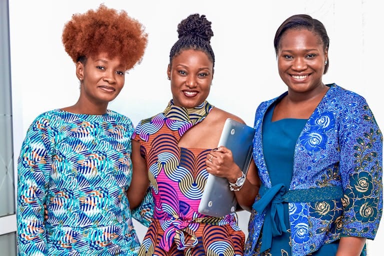 Three diverse businesswomen smiling in vibrant attire, showing teamwork indoors.