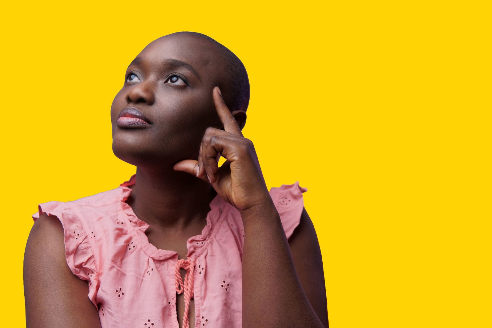 Bald African American woman in pink top looking contemplative against bright yellow background.